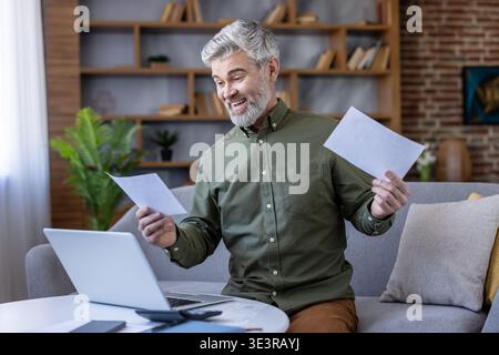 Senior man smiling excitedly while reviewing paperwork and using a laptop at a home office setup, celebrating financial success and managing bills online Stock Photo