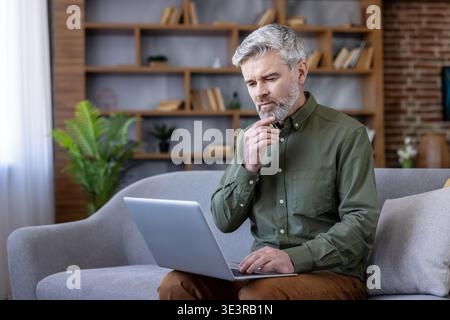 Thoughtful middle-aged man with grey hair and beard sitting on a sofa, concentrating while using his laptop for remote work or online studying in a comfortable home office setting Stock Photo