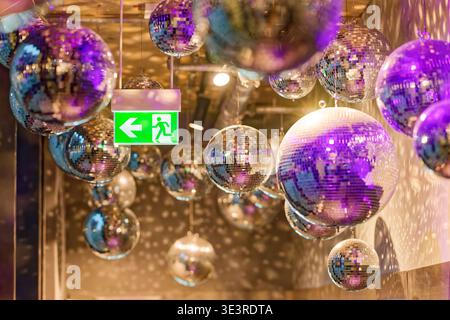 Multiple reflective disco balls suspended from the ceiling create colorful light patterns around a glowing green emergency exit sign in an indoor sett Stock Photo