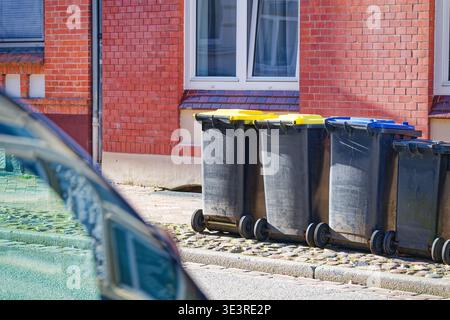 Several black garbage bins with yellow and blue lids are lined up on a cobblestone sidewalk next to a red brick building with white framed windows. Stock Photo