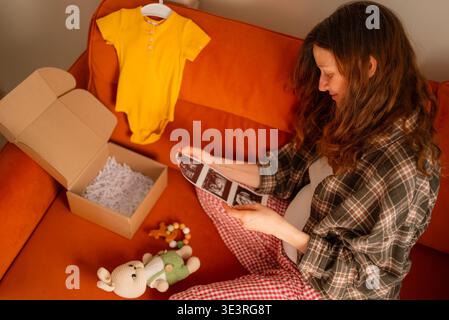 Pregnant woman anticipating motherhood, holding sonogram images while sitting on a couch near baby clothes, a cardboard box, and a knitted toy, preparing for new life Stock Photo