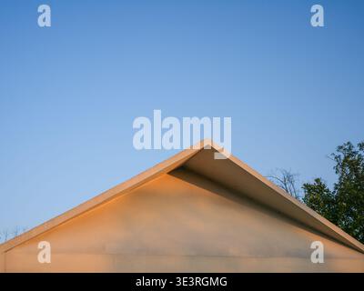 A minimalist view of a triangular building roof peak illuminated by warm golden sunlight against a vast clear blue sky. The scene represents architect Stock Photo