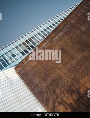 An abstract low angle view of a modern geometric building facade featuring contrasting white panels, glass windows, and rusty perforated metal. The sc Stock Photo