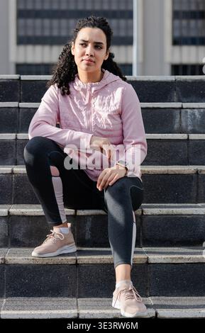 Woman sitting on concrete steps outside building wearing pink jacket leggings smartwatch, shoes Stock Photo