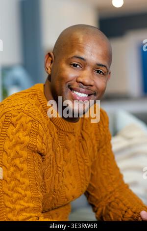 African American man wearing mustard sweater sitting on sofa at home, smiling at camera Stock Photo