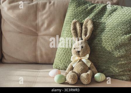 Cozy Easter scene featuring plush bunny, decorative pastel eggs arranged on sofa with textured pillows. Stock Photo