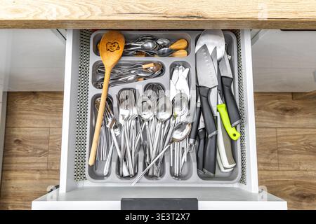 Open cutlery drawer, knives, forks, spoons, teaspoons, tableware placed in the drawer, new practical kitchen furniture with white drawers, wooden furn Stock Photo