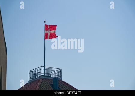 A red flag with a white cross is flying on a flagpole mounted on a rooftop with a railing, set against a clear blue sky background. Stock Photo