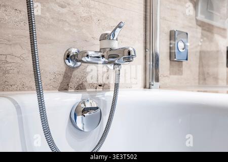 Close up of a sleek silver single-handle mixer tap on a white bathtub with a flexible shower hose against a light beige stone-textured tile wall Stock Photo