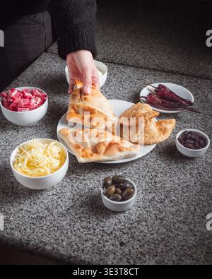 Hand holds freshly baked bread with golden crust. Bowl of cheese, olives, pickled onions, and red peppers surround plate. Simple, rustic meal setup on Stock Photo