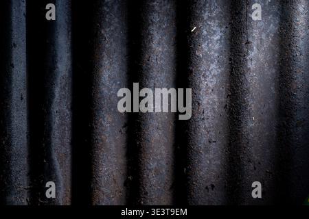 Macro shot of old, dirty, and heavily rusted dark corrugated iron showing vertical waves and weathered texture under high contrast side lighting. Stock Photo
