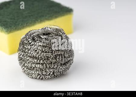 A close-up view of a shiny stainless steel wire wool scrubber ball positioned in front of a yellow foam and green abrasive sponge on a white backgroun Stock Photo