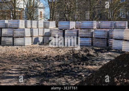 Pallets of concrete paving blocks stacked on a construction site, with muddy terrain and residential buildings awaiting reconstruction work Stock Photo