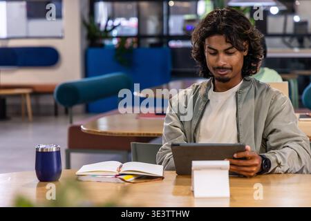 Indian man sitting at wooden table on right, using tablet on stand, notebook, notes, copy space Stock Photo