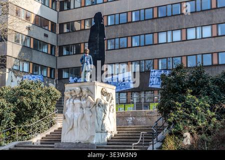 A monument featuring a relief sculpture on the plinth and a black silhouette on the stele, set against a GDR prefabricated block of flats. On the white marble plinth with the relief sculpture stands a small, clothed figure in Rococo costume, with a larger-than-life black silhouette behind it; in the background, historical photographs adorn the façade. Leipzig, Saxony, Germany Stock Photo