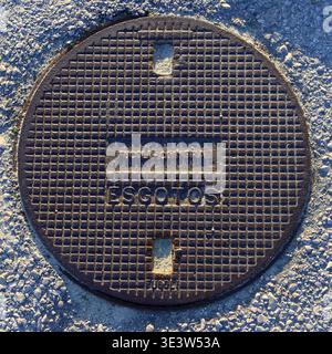Costa Nova, Portugal - March 12, 2026: Round rusted manhole cover with grid pattern and “FUCAL - OMBRA” and “ESGOTOS” text embedded in gravel ground Stock Photo