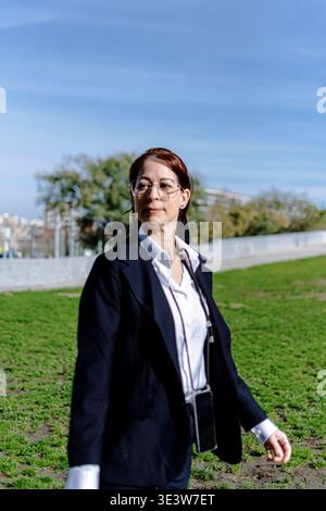 Businesswoman with glasses and suit jacket walking confidently through a grassy park, looking away from the camera, carrying a smartphone on a lanyard Stock Photo