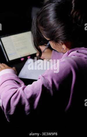 Woman studies late at night, focused on laptop screen. Her purple sweater blends with soft ambient glow. Dark room highlights her concentration and qu Stock Photo