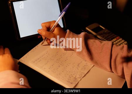 Soft light glows on notebook and pen. Hand writes carefully, focused and calm. Digital tablet displays blank screen nearby. Calculator rests quietly b Stock Photo