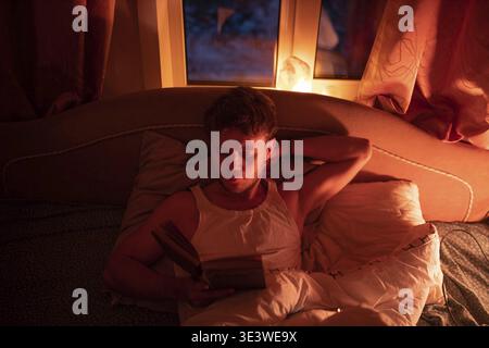 Man relaxed in bed reading a book by soft, warm light near a window Stock Photo