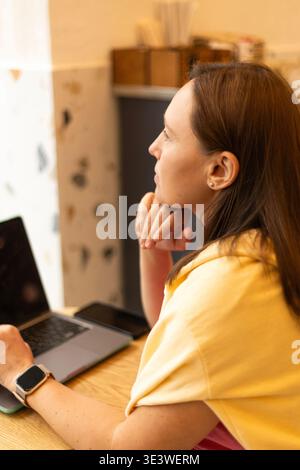 A close-up of a woman resting her head on her hand, thoughtfully gazing while sitting in front of a laptop at a cafe, capturing a moment of deep refle Stock Photo