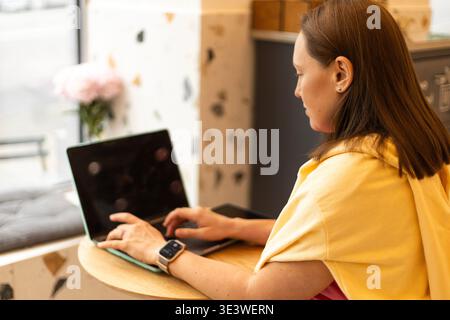 A woman types on her laptop by the window in a cozy cafe. The setting is bright and inviting, with a focus on productivity and relaxation in a comfort Stock Photo