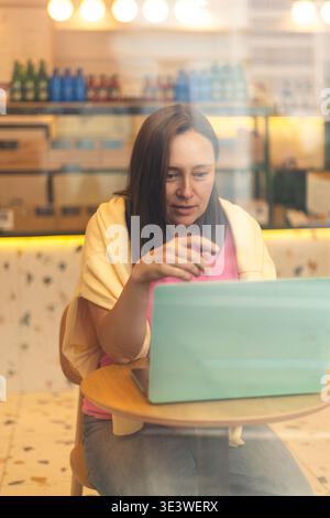 A young woman engages in an online video call on her laptop while seated comfortably by a window in a cozy cafe, surrounded by a warm and inviting atm Stock Photo