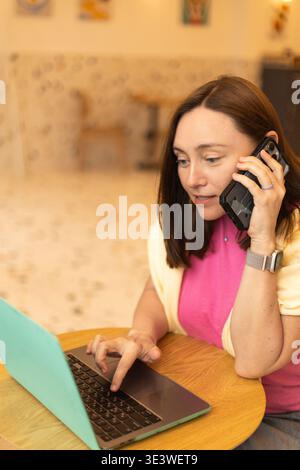A freelancer woman sits in a cafe, using her laptop and talking on the phone. She efficiently manages her tasks, illustrating a modern, mobile work st Stock Photo