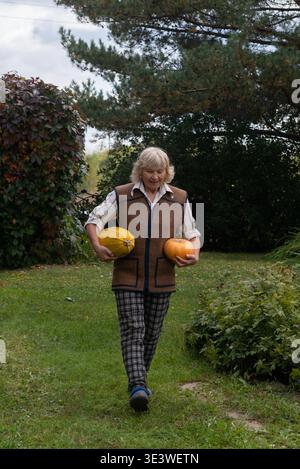 An elderly woman enjoys a walk in her lush garden, carrying two freshly harvested pumpkins. The vibrant greenery and colorful foliage create a serene, Stock Photo
