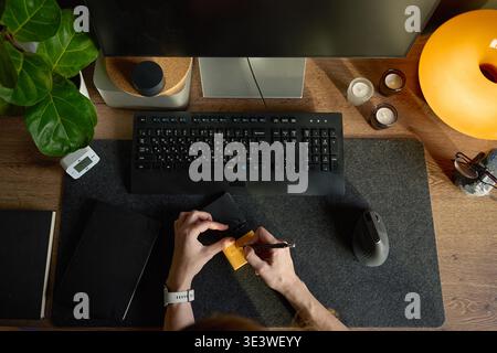 Top view of hands writing on sticky note at desk with monitor, keyboard, mouse and plant Stock Photo