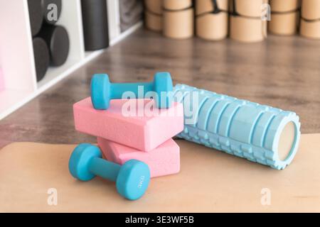 A set of blue dumbbells, pink yoga blocks, and a textured foam roller are neatly arranged on a yoga mat in a gym, promoting fitness and well-being in Stock Photo