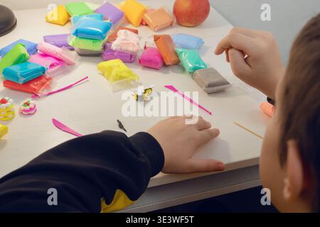 A child engaged in creative crafting, using colorful modeling clay on a white table. Various clay packs and small sculpting tools are scattered around Stock Photo