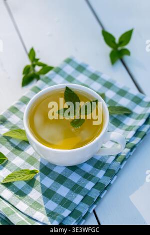 A soothing cup of homegrown mint tea placed on a green checkered cloth, surrounded by fresh mint leaves on a wooden table, capturing a moment of tranq Stock Photo
