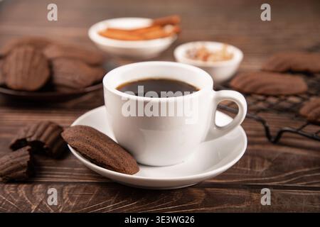 A steaming cup of coffee accompanies chocolate madeleines shaped like seashells, resting on a rustic wooden table, creating an inviting and warm morni Stock Photo