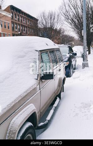 Snow-covered vehicles line a residential street in an urban setting, showcasing the aftermath of a winter storm. Trees and buildings are visible under Stock Photo
