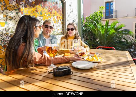 Three friends sitting at an outdoor table, sharing tapas and drinks. They are enjoying each other's company, socializing and making authentic connecti Stock Photo