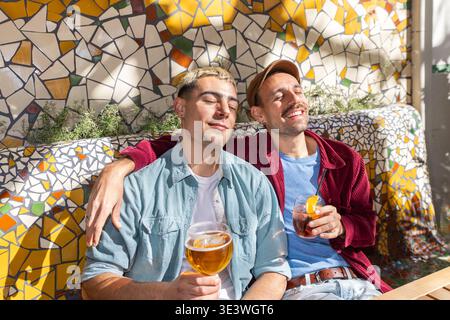Two friends relax and enjoy drinks together, basking in the warm sunlight. Their smiles and casual attire reflect the joy of socialising, connection, Stock Photo