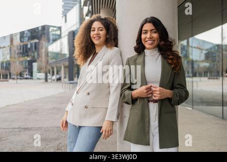 Two confident women in professional attire lean against a column in an urban environment, smiling warmly. Their business style reflects teamwork and f Stock Photo
