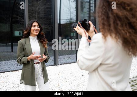 Two professional women collaborate outdoors, showcasing teamwork in a business environment. One holds a camera while capturing the other speaking conf Stock Photo