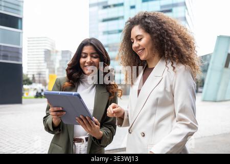 Two smiling women in business attire stand outside a modern building, collaborating on a tablet. Their teamwork and friendship reflect a positive work Stock Photo
