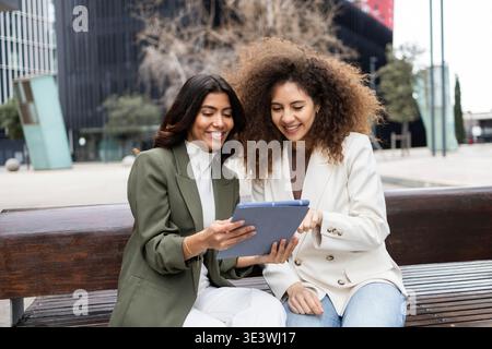 Two professional women sitting on a bench, smiling and collaborating on a business project using a tablet. They demonstrate teamwork and friendship in Stock Photo