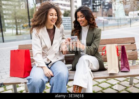 Two women sitting on a bench and enjoying a day out with shopping bags. They are sharing a pleasant moment together, possibly discussing purchases or Stock Photo