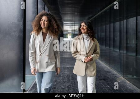 Two women walk confidently through a sleek, modern hallway, embodying professionalism and friendship. Their smiles reflect a strong bond as they navig Stock Photo