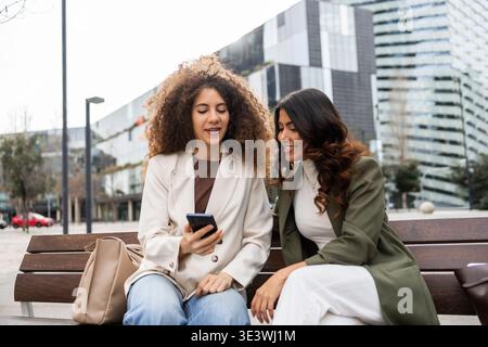 Two women sitting on a bench outdoors, discussing ideas with a smartphone in hand. Dressed in business attire, they symbolize teamwork and friendship Stock Photo