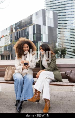 Two women sitting on a bench, laughing and looking at a smartphone, taking a break from work in an urban business district, showcasing friendship and Stock Photo