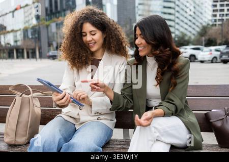 Two businesswomen sitting on a bench in the city, engaged in a lively discussion while looking at a tablet. They are friends collaborating on a projec Stock Photo