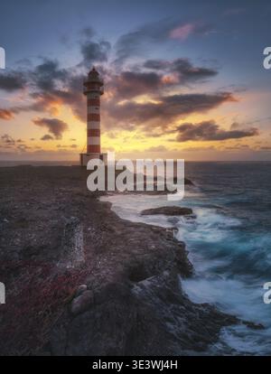 The Punta Sardina Lighthouse stands majestically against a vibrant sunset. Waves crash against the rocky coastline, while dramatic clouds enhance the Stock Photo