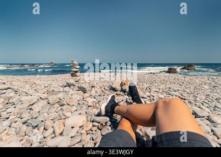 A person relaxes on the pebbled shore of Campiechos Beach in Asturias, enjoying the serene ocean view with stacked stones nearby, under a clear blue s Stock Photo