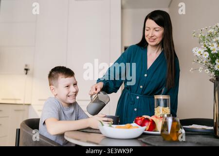 A joyful mother pours a drink for her smiling son at the breakfast table, surrounded by fruits and snacks, capturing a warm family moment in a modern Stock Photo