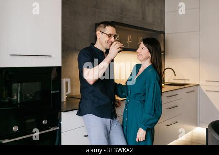 A couple shares a joyful moment while enjoying a drink in a sleek, modern kitchen. The warm atmosphere highlights their connection and the stylish int Stock Photo